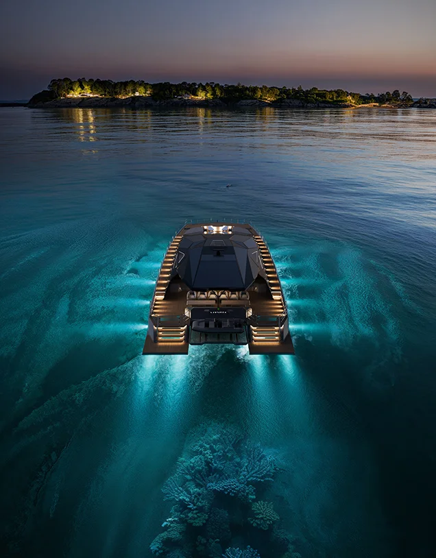 Rear aerial night view of the LANIAKEA luxury power catamaran cruising over illuminated turquoise waters toward a private island