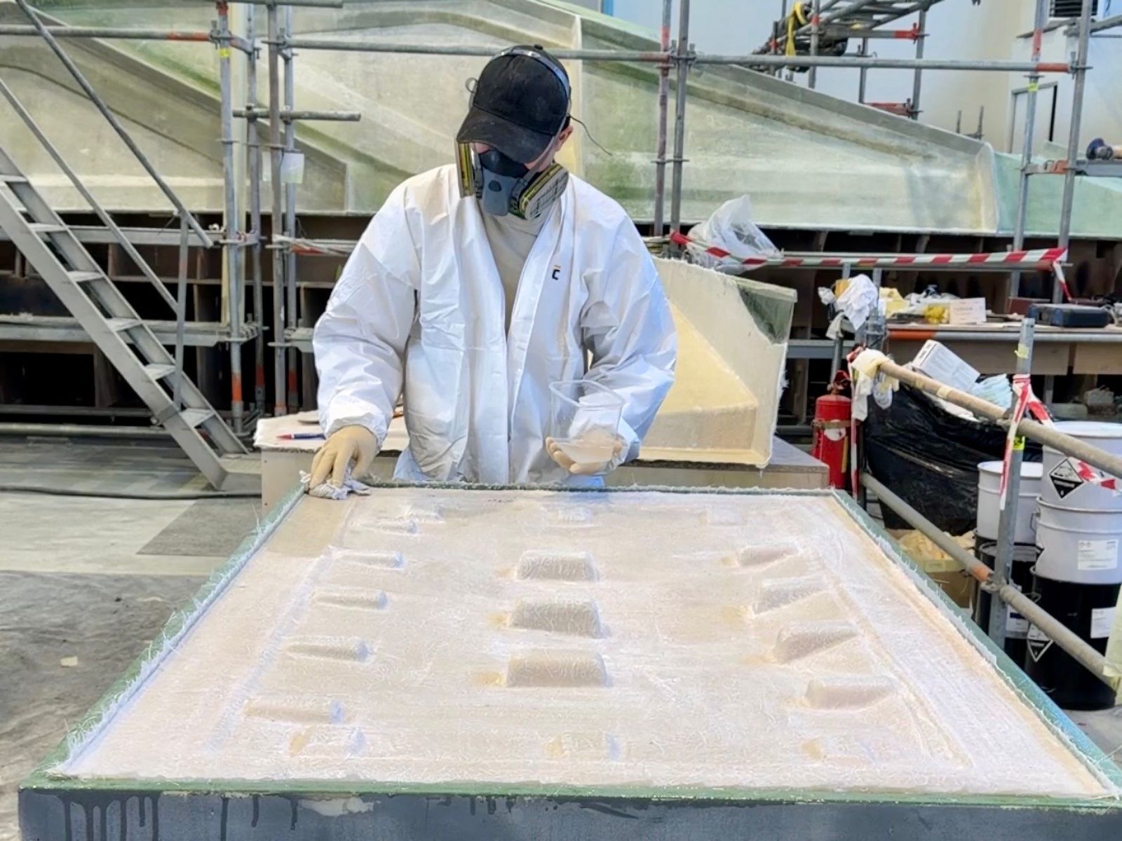 Worker in protective gear applying resin to a fiberglass mold in a shipyard